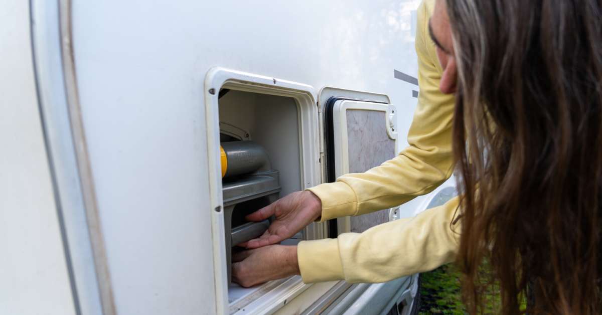 A woman working on the water tank of her RV. She is currently pulling it out of the side of the vehicle.