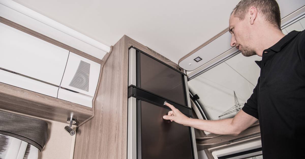 A man reaching out with his hand and pointing at an RV fridge. The refrigerator is built into the wall of the RV.