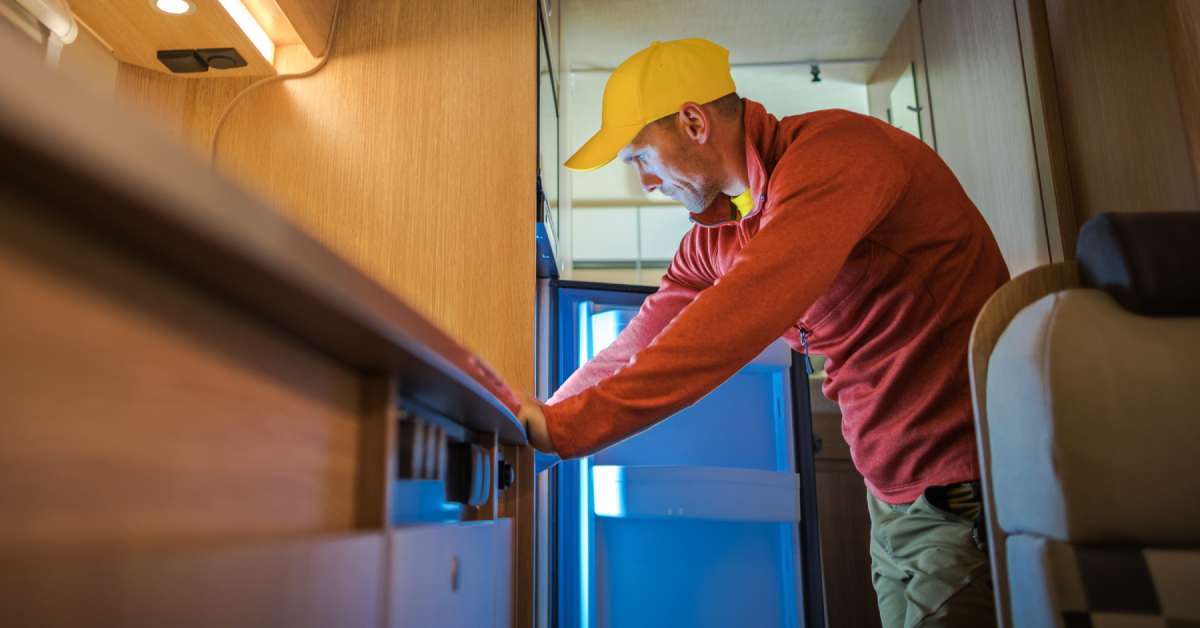 A man with a red coat and yellow baseball cap looking inside of his RV refrigerator with both hands on the counter.